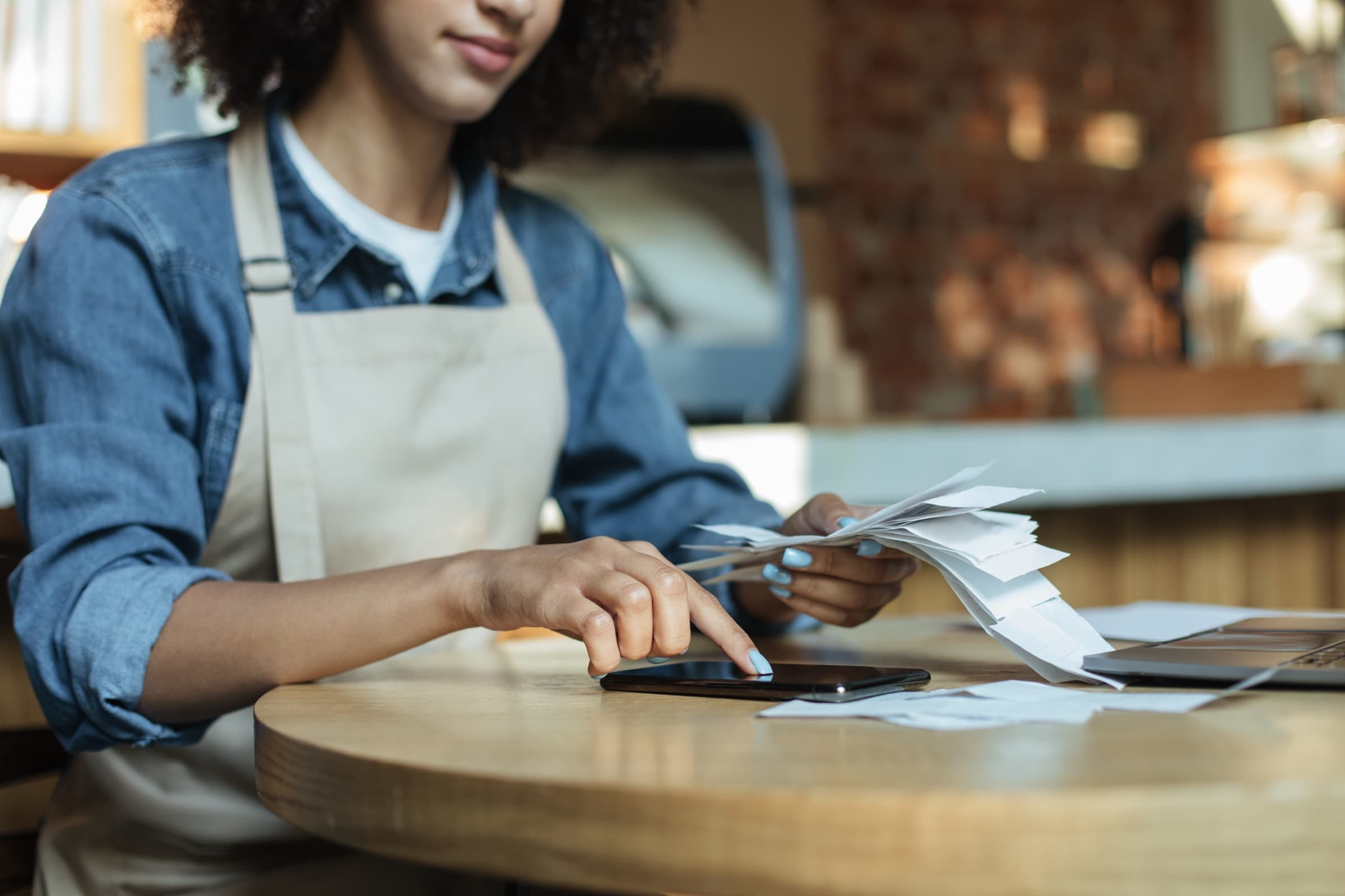 A café owner in an apron at a wooden table, working through a handful of receipts